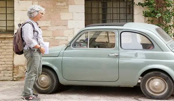 Woman on a hike, looking at a small grey car parked in an Italian street in front of a natural stone building.