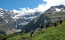 People walking down a mountain track in the French Pyrenees. A wide mountainous landscape with snowcapped mountains, a blue sky and a high waterfall in the distance