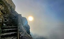 Sun shinging through mist next to steps on the side of a mountain leading up to the ruins of a castle